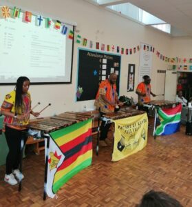 Three people wearing national dress, playing African percussion instruments.