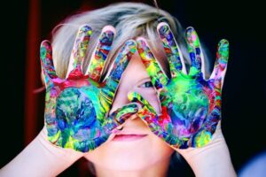 Child holding their hands in front of their face. Their hands are covered in bright paint.